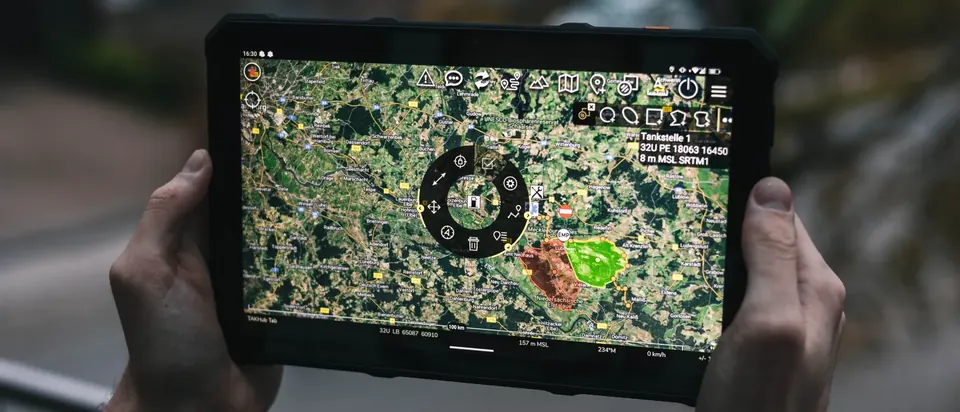 Zwei Hände halten ein robustes Tablet mit ATAK. Auf der Satellitenkarte sind zwei Einsatzabschnitte rot und grün eingezeichnet, dazu ein Marker mit Koordinate.