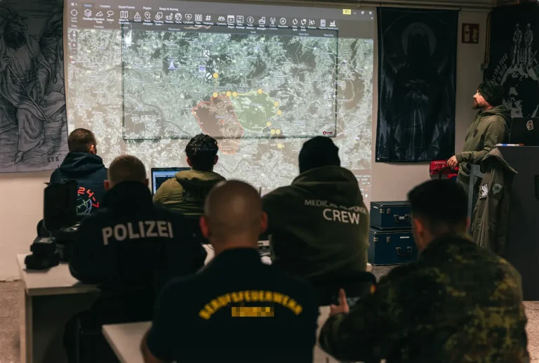 Воркшоп з ATAK: учасники з поліції, пожежної охорони, медичної служби та Бундесверу стежать за картою обстановки ATAK на проєкційному екрані, праворуч стоїть тренер.